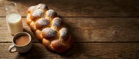 Fresh braided challah bread with powdered sugar on rustic wooden table alongside glass of milk and coffee cup for bakery and breakfast concepts.