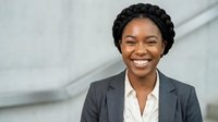 Professional African American businesswoman with braided hair smiling confidently in corporate suit standing outdoors near modern building architecture.