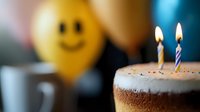 Birthday cake with lit candles in foreground and yellow smiley balloon in background, creating festive celebration atmosphere with soft bokeh lighting effect.