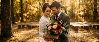 Romantic wedding couple embracing in golden autumn park with fall foliage. Bride in lace dress holds burgundy bouquet while groom in suit tenderly holds her.