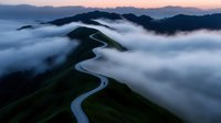 Winding river cuts through green mountain ridges emerging from sea of white clouds at dawn with purple sky and distant peaks creating mystical landscape.