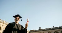 Middle-aged man with dark skin tone wearing black academic regalia and mortarboard cap, holding diploma certificate outdoors near university building on graduation day.