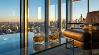 Luxury penthouse interior with whiskey glasses on table overlooking city skyline at sunset through floor-to-ceiling windows creating elegant urban lifestyle scene.