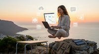Young professional woman working remotely on laptop with digital analytics dashboard overlooking scenic ocean sunset from mountain cliff location.
