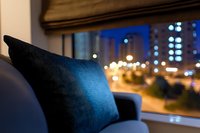 Cozy blue velvet cushion on sofa near window with blurred night city lights and buildings in background creating peaceful evening atmosphere.