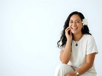 Young woman with long black hair and South Asian ethnicity wearing casual white t-shirt, speaking on mobile phone with joyful expression, white flower accessory in hair, isolated on white background for design use.