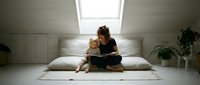 Young mother reading book with toddler daughter on bed in bright minimalist bedroom. Family bonding time, early childhood education and parenting moments.