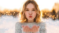 Young woman with blonde wavy hair blowing kiss in winter setting with bokeh lights, wearing cozy sweater. Perfect for seasonal greeting cards.