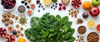 Fresh leafy greens surrounded by colorful superfoods including berries, nuts, seeds and citrus fruits on white background for healthy eating concepts.