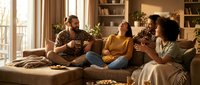 Friends sitting on couch having conversation in cozy living room with warm lighting, shelves, and plants. Use for lifestyle, friendship, home decor, and social gathering content.