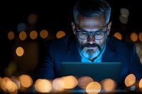 Mature Caucasian businessman with gray beard and glasses working late on digital tablet, illuminated by blue screen light against dark background with bokeh.