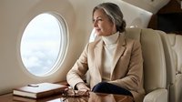 Elegant mature businesswoman in beige suit relaxing in luxury private jet cabin, looking through window during flight for corporate travel concepts.