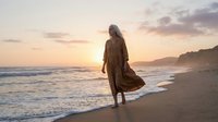 Woman in flowing brown dress walking along ocean beach at golden sunset with waves and cliffs creating peaceful coastal scene for wellness and travel concepts.