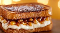 Grilled sandwich with melted cream cheese and caramelized nuts on golden toasted bread, dusted with powdered sugar against warm bokeh background.