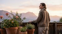 Woman in casual brown cardigan standing on balcony with potted plants, gazing at mountain landscape during golden hour sunset. Lifestyle and travel concept.