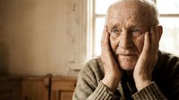 Elderly Caucasian man with hands on face showing contemplative expression in warm indoor setting with natural window light for healthcare concepts.