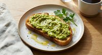 Avocado toast on sourdough bread with herbs and olive oil drizzle served on ceramic plate with coffee cup on wooden table for healthy breakfast meal.