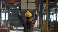 Young Asian female warehouse worker wearing yellow hard hat and plaid shirt lifting cardboard boxes overhead in industrial storage facility. Safety at workplace.