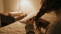 Young woman with long dark hair in cozy knit sweater holding warm cup in softly lit bedroom creating peaceful morning or evening atmosphere.