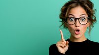 Young Caucasian woman with glasses showing surprised expression, raised index finger against turquoise background, messy bun hairstyle wearing black top, copy space.