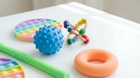 Colorful sensory toys including textured ball, fidget spinner, pop-it toy and rubber ring arranged on white surface for child development and play.