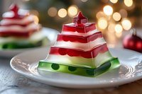 Festive layered jello dessert in red, white and green stripes shaped like Christmas tree on white plate with bokeh lights background creates holiday atmosphere.