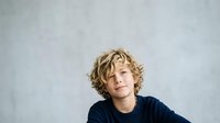 Young boy with curly blonde hair wearing dark navy shirt, looking upward thoughtfully against neutral gray background, suitable for educational, parenting, and lifestyle content.