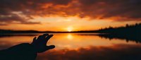 Hand silhouette reaching toward golden sunset over calm lake with perfect reflection. Dramatic orange sky creates peaceful moment for meditation and hope concepts.