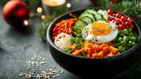 Fresh colorful buddha bowl with fried egg, carrots, cucumber, red currants and microgreens served in black stone bowl against dark background with holiday lights.