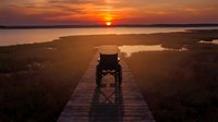 Wheelchair user on wooden boardwalk at sunset overlooking wetlands and water, accessibility in nature, outdoor recreation and inclusive travel.