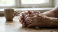 Elderly hands clasped together on wooden table beside coffee mug in soft natural light, conveying prayer, contemplation, and peaceful moments of reflection.
