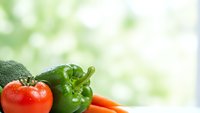 Fresh organic vegetables including red tomato, green bell pepper, broccoli and carrots arranged on white surface with soft natural lighting and bokeh background.