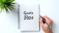 Hand holding pen writing goals for 2026 in spiral notebook on white background with green plant, representing future planning and personal development.