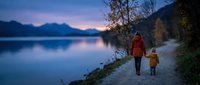 Mother and child walking together along peaceful lake shore during sunset with mountains in background. Family bonding time in nature during golden hour twilight.