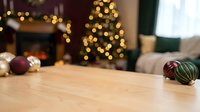Wooden table surface with Christmas ornaments in cozy living room with fireplace and decorated tree with warm bokeh lights in background.