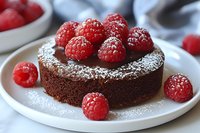 Rich chocolate cake topped with fresh red raspberries and dusted with powdered sugar on white plate, close-up view of homemade dessert with vibrant berries.