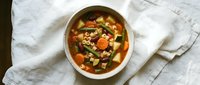 Hearty vegetable soup with carrots, green beans and pasta in savory broth served in ceramic bowl on white linen napkin for healthy comfort food recipes.