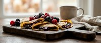 Delicious homemade pancakes topped with fresh mixed berries and syrup served on rustic wooden board with coffee mug in cozy kitchen setting for breakfast.