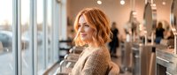 Young redhead woman smiling in modern hair salon with stylists working in background. Professional beauty and haircare service concept.
