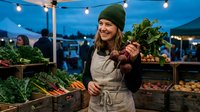 Young Caucasian female farmer in apron and beanie holding fresh leafy greens at evening farmers market with string lights and organic produce displays.