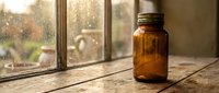 Empty amber glass jar on rustic wooden windowsill with warm sunlight streaming through rain-spotted window creating cozy atmospheric lighting for home decor.