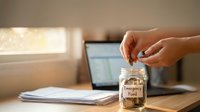 Person saving coins in emergency fund jar on wooden desk with laptop and documents for financial planning and budgeting preparation.
