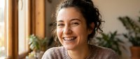 Happy young woman with curly hair smiling warmly in cozy home interior with natural lighting and plants creating welcoming atmosphere for lifestyle content.
