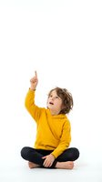 Curious young boy in yellow hoodie pointing up with finger while sitting cross-legged, isolated on white background for educational and childhood concepts.