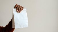 Hand holding blank white paper bag against neutral background for packaging, branding, and retail mockup purposes.