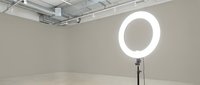 Glowing ring light on a stand in a minimalist studio space with neutral beige walls, concrete floor, and ceiling track lighting.