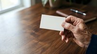 Mature man with medium skin tone holding blank white business card at wooden desk with plant and window in background, suitable for corporate identity and professional networking concepts.