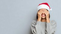 Young woman in Santa hat covering eyes with hands while smiling excitedly against gray background, expressing holiday surprise and joy.