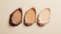Makeup foundation smears in different skin tone shades from dark to light on beige background, showing cosmetic texture and color range for beauty products.