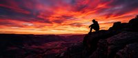 Silhouetted male climber perched on rocky summit against dramatic fiery sunset sky with vibrant orange and red clouds, suitable for adventure, motivation and outdoor sports marketing.